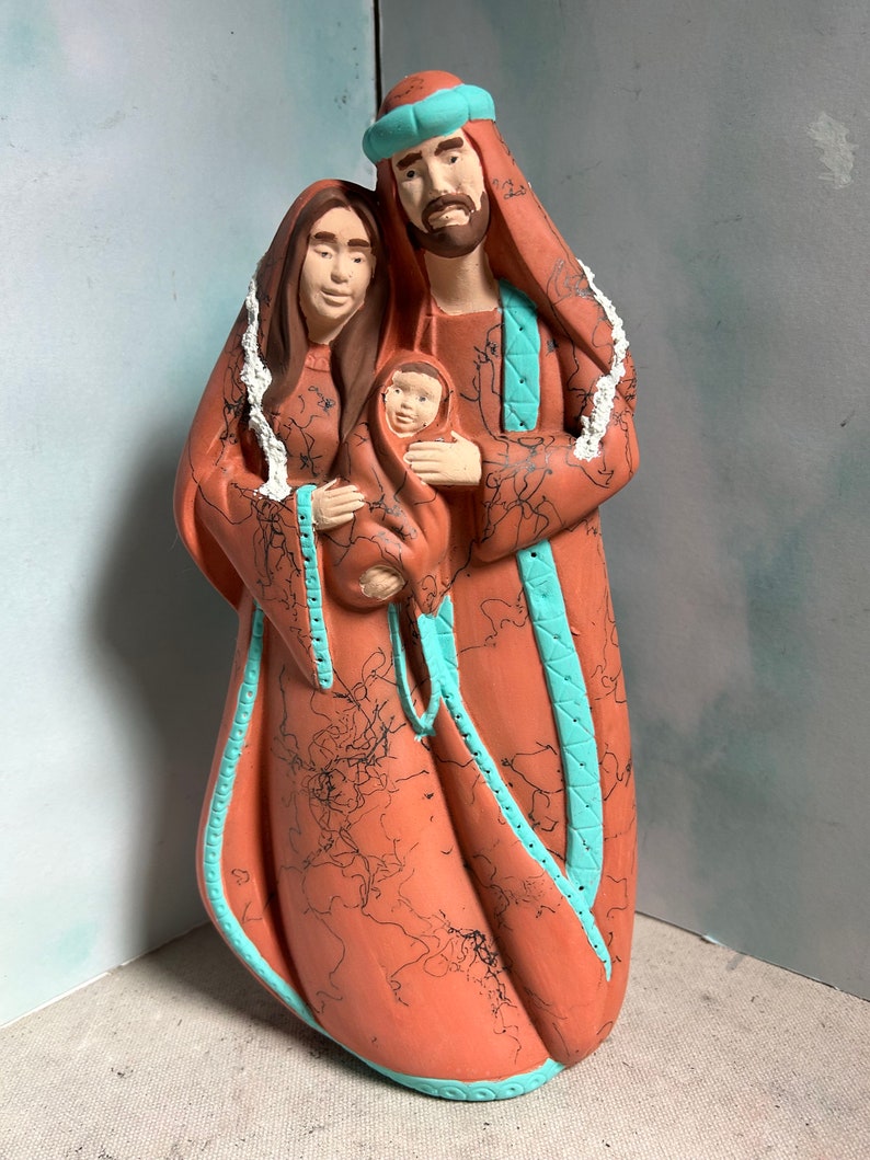 Puede incluir: Una figurita de cer&aacute;mica de la Sagrada Familia. Las figuras est&aacute;n pintadas en tonos marrones y rojos, con detalles turquesa. La madre sostiene un beb&eacute; en sus brazos. El padre est&aacute; de pie detr&aacute;s de ellos, con su brazo alrededor de la madre.