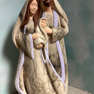 Puede incluir: Una figurita de cer&aacute;mica de la Sagrada Familia. Las figuras est&aacute;n pintadas en beige y marr&oacute;n con acentos morados. La madre sostiene un beb&eacute; envuelto en un pa&ntilde;o blanco. El padre est&aacute; de pie detr&aacute;s de la madre con su brazo alrededor de ella.