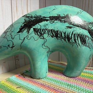May include: A turquoise ceramic bear sculpture with black abstract markings. The bear has a smooth, rounded form with a large body and two legs. The sculpture is placed on a colorful, striped textile.