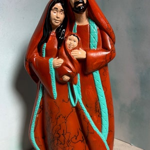 Puede incluir: Una figurita de cer&aacute;mica de la Sagrada Familia. Las figuras est&aacute;n pintadas en tonos rojos y turquesa. La madre sostiene un beb&eacute; en sus brazos. El padre est&aacute; de pie detr&aacute;s de ellos con su brazo alrededor de la madre.
