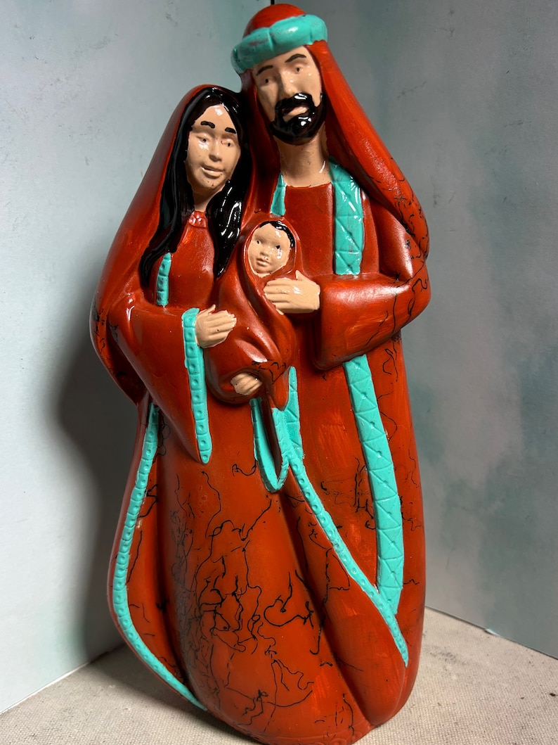 Puede incluir: Una figurita de cer&aacute;mica de la Sagrada Familia. Las figuras est&aacute;n pintadas en tonos rojos y turquesa. La madre sostiene un beb&eacute; en sus brazos. El padre est&aacute; de pie detr&aacute;s de ellos con su brazo alrededor de la madre.