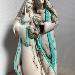 Puede incluir: Una escultura de cer&aacute;mica blanca de la Sagrada Familia. Las figuras est&aacute;n representadas en un estilo tradicional, con Mar&iacute;a sosteniendo al ni&ntilde;o Jes&uacute;s y Jos&eacute; de pie a su lado. La escultura est&aacute; decorada con l&iacute;neas negras y acentos turquesa.
