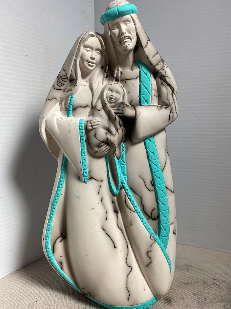 Puede incluir: Una escultura de cer&aacute;mica blanca de la Sagrada Familia. Las figuras est&aacute;n representadas en un estilo tradicional, con Mar&iacute;a sosteniendo al ni&ntilde;o Jes&uacute;s y Jos&eacute; de pie a su lado. La escultura est&aacute; decorada con l&iacute;neas negras y acentos turquesa.