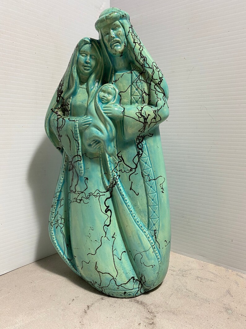 Puede incluir: Una figurita de cer&aacute;mica de la Sagrada Familia, que representa a Mar&iacute;a, Jos&eacute; y el ni&ntilde;o Jes&uacute;s. La figurita est&aacute; pintada en tonos azules y verdes con acentos negros. Las figuras est&aacute;n de pie juntas, con Mar&iacute;a sosteniendo al ni&ntilde;o Jes&uacute;s en sus brazos.