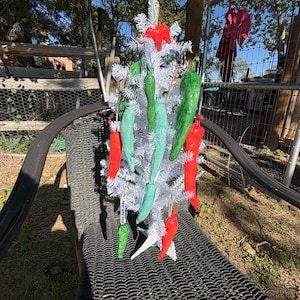 Puede incluir: Un pequeño árbol artificial blanco decorado con múltiples adornos colgantes en forma de chiles en tonos rojo brillante, verde claro y verde oscuro. El árbol tiene agujas blancas y se apoya sobre una base blanca de cuatro puntas.