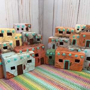 May include: Two multi-level ceramic structures resembling pueblo-style buildings. Each piece features rectangular windows, brown doors, and turquoise accents. The surfaces have a textured, mottled finish with earthy tones of tan, orange, and blue.