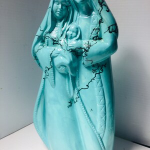 Puede incluir: Una figurita de cer&aacute;mica azul turquesa de la Sagrada Familia. Las figuras est&aacute;n vestidas con t&uacute;nicas con detalles de craquelado negro. La madre sostiene al ni&ntilde;o Jes&uacute;s.