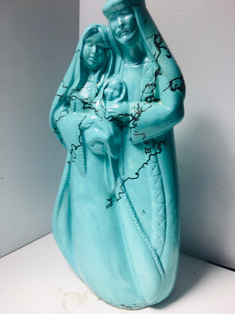 Puede incluir: Una figurita de cer&aacute;mica azul turquesa de la Sagrada Familia. Las figuras est&aacute;n vestidas con t&uacute;nicas con detalles de craquelado negro. La madre sostiene al ni&ntilde;o Jes&uacute;s.