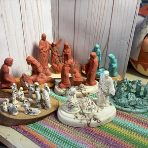 Puede incluir: Una colección de varias figuras de belén hechas de arcilla con diversos acabados, incluyendo terracota, turquesa y blanco con patrones de grietas negras. Las figuras están dispuestas sobre rodajas de madera y bases circulares.