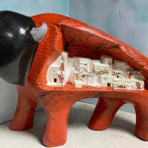 Puede incluir: Una escultura de cerámica roja de un bisonte con un interior tallado que representa un pueblo blanco. El bisonte tiene cuernos negros y una cabeza negra.