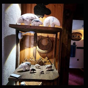 May include: A wooden shelf displays a collection of polar bear sculptures. The bears vary in size and are white with grey markings. The shelf is part of a rustic wooden structure, with a woven basket and mountain-shaped decor.