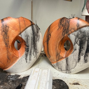 以下が含まれることがあります： ユニークなデザインのセラミック製の壺が2つ。壺は薄い茶色で黒い線が入り、白い部分には黒い線が描かれている。壺はドーナツ型で、中央に穴が開いている。