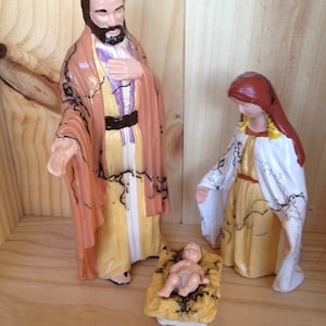 Puede incluir: Un conjunto de figuras de nacimiento que incluye una figura masculina de pie con túnica color melocotón y amarilla, una figura femenina de pie con túnica blanca y amarilla con velo rojo, y un bebé en un pesebre de textura amarilla.
