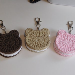 May include: Three crocheted bear-shaped keychains in brown, tan, and pink. Each keychain has a white layer in the middle, resembling a sandwich cookie. The keychains have silver-colored clasps.
