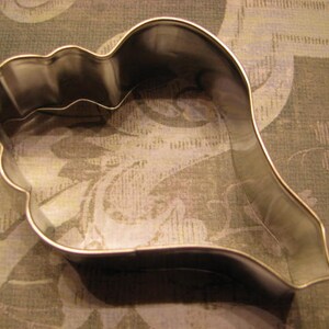 May include: A silver metal cookie cutter shaped like a seashell. The cutter has a smooth, rounded surface and a slightly curved shape.