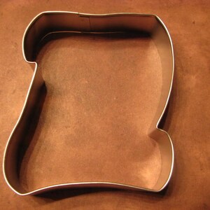 May include: A silver metal cookie cutter shaped like a rolled up scroll or parchment paper.