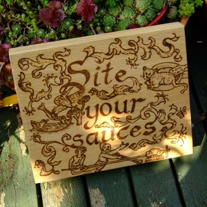 Puede incluir: Un letrero de madera con las palabras "Site your sauces" quemadas en la superficie. El letrero está decorado con intrincados diseños florales y una pequeña ilustración de un animal.
