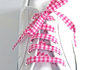 May include: White canvas sneaker with pink and white gingham laces. The laces are tied in a bow. The shoe has a white rubber toe cap and a blue trim around the sole. The shoe is on a white background.