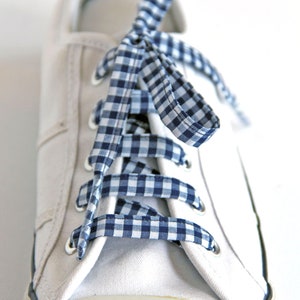 May include: Close-up of a white canvas sneaker with blue and white gingham ribbon laces. The toe cap is off-white, and the sole has a black trim. The laces are tied in a bow.