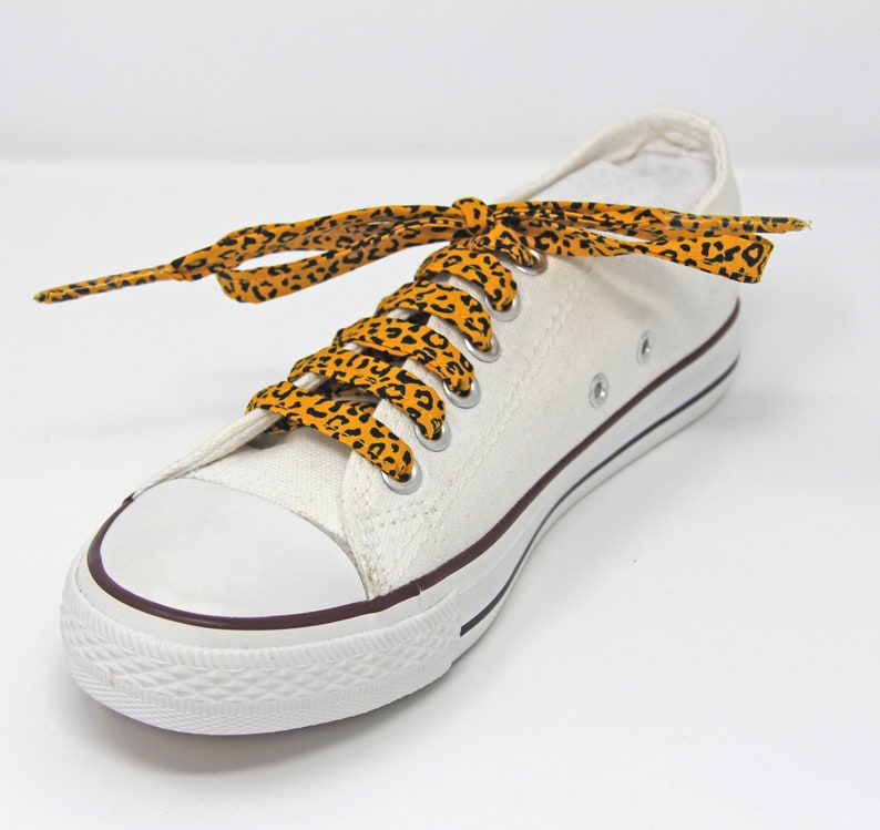 K&ouml;nnte beinhalten: Wei&szlig;e Canvas-Sneaker mit niedrigem Schaft und orangefarbenen und schwarzen Schn&uuml;rsenkeln mit Leopardenmuster.