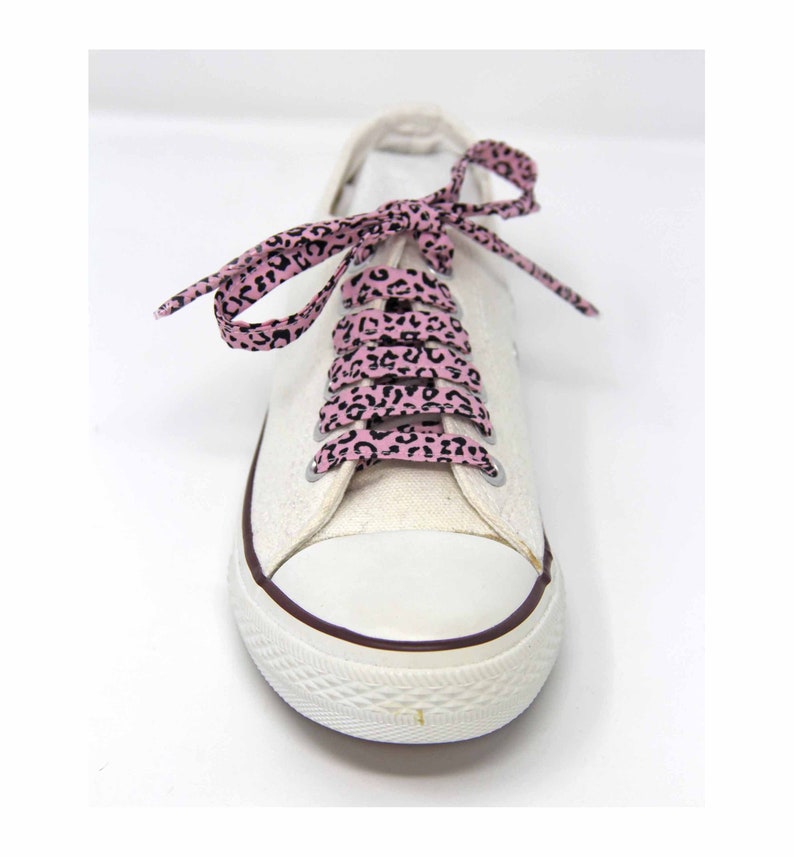 K&ouml;nnte beinhalten: Wei&szlig;e Canvas-Sneaker mit pinkfarbenen und schwarzen Schn&uuml;rsenkeln im Leopardenmuster. Die Schn&uuml;rsenkel sind oben an der Schuhspitze zu einer Schleife gebunden.