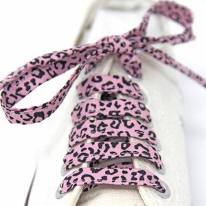 K&ouml;nnte beinhalten: Rosa Schn&uuml;rsenkel mit einem schwarzen Leopardenmuster. Die Schn&uuml;rsenkel sind an einem Paar wei&szlig;en Sneakers gebunden.