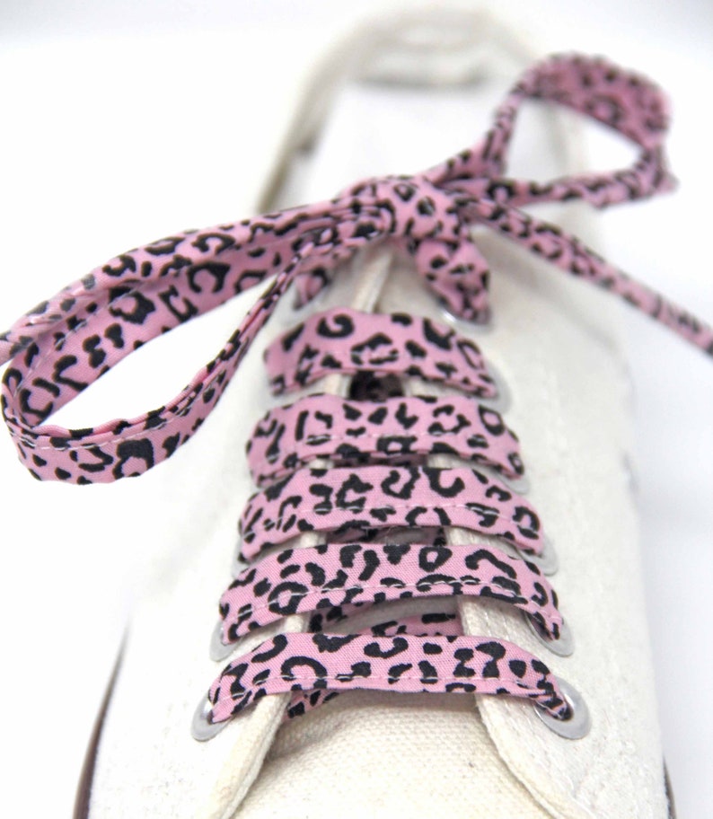 K&ouml;nnte beinhalten: Rosa Schn&uuml;rsenkel mit einem schwarzen Leopardenmuster. Die Schn&uuml;rsenkel sind an einem Paar wei&szlig;en Sneakers gebunden.