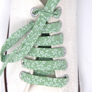 May include: White canvas sneakers with green and white floral patterned shoelaces. The laces are tied in a criss-cross pattern.