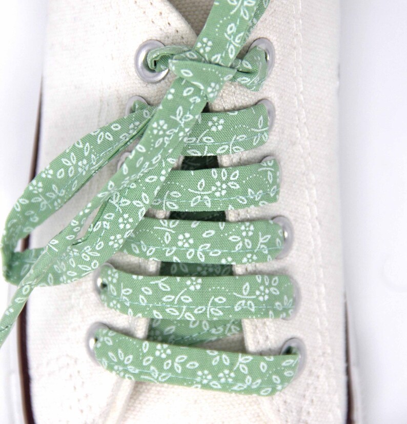 May include: White canvas sneakers with green and white floral patterned shoelaces. The laces are tied in a criss-cross pattern.