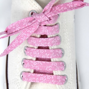 May include: Pink shoelaces with a white floral pattern are tied on a pair of white canvas shoes.