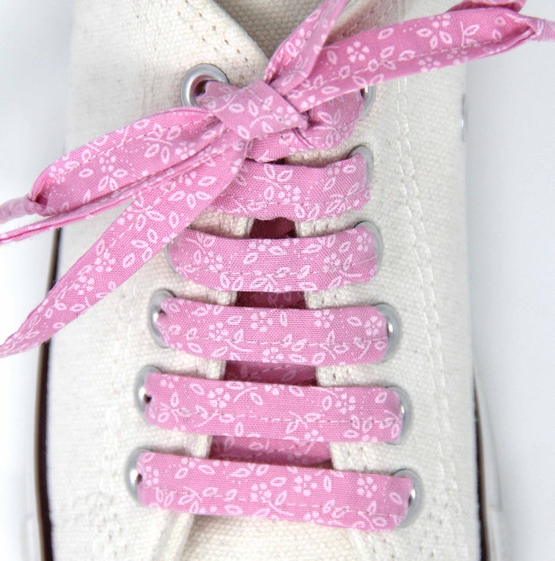 May include: Pink shoelaces with a white floral pattern are tied on a pair of white canvas shoes.
