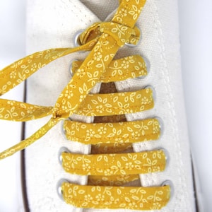 May include: White canvas shoes with yellow floral patterned laces tied in a criss-cross pattern.