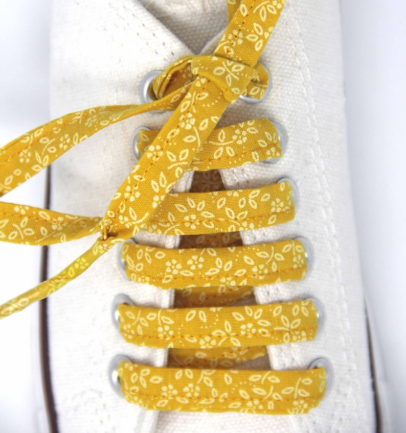 May include: White canvas shoes with yellow floral patterned laces tied in a criss-cross pattern.