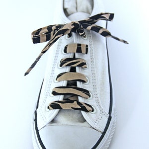 K&ouml;nnte beinhalten: Wei&szlig;e Canvas-Sneaker mit schwarzen und braunen Schn&uuml;rsenkeln im Zebra-Muster. Die Schn&uuml;rsenkel sind im Kreuzmuster gebunden.