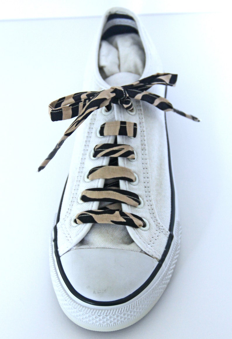 K&ouml;nnte beinhalten: Wei&szlig;e Canvas-Sneaker mit schwarzen und braunen Schn&uuml;rsenkeln im Zebra-Muster. Die Schn&uuml;rsenkel sind im Kreuzmuster gebunden.