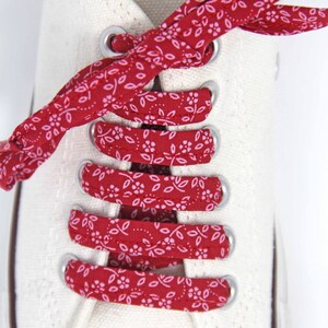 May include: Red and white patterned shoelaces are tied in a criss-cross pattern on a pair of white canvas shoes.