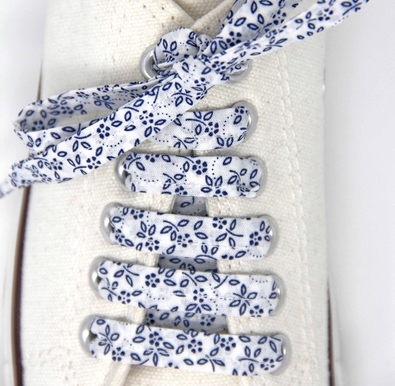 May include: White canvas shoes with blue and white patterned shoelaces. The shoelaces are tied in a bow at the top of the shoe.