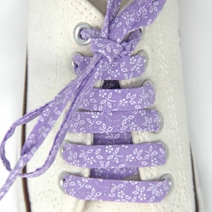 May include: White canvas sneakers with purple floral shoelaces tied in a criss-cross pattern.