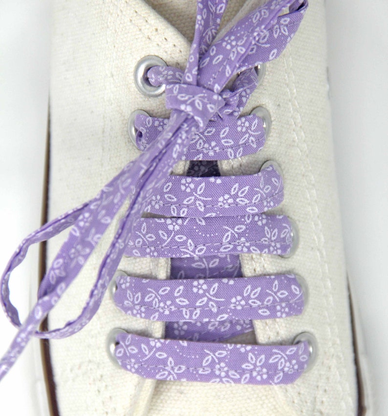 May include: White canvas sneakers with purple floral shoelaces tied in a criss-cross pattern.