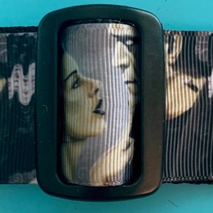 May include: Black and white patterned fabric belt with a black buckle. The fabric features a repeated image of a couple, a man and a woman, with a gothic horror theme. The woman has black hair and a white scar on her forehead. The man has black hair and a scar on his forehead.