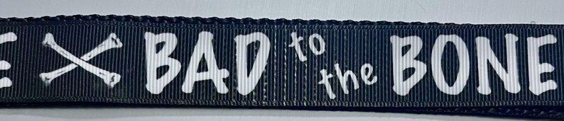 May include: Black dog leash with white text and crossed bones. The text reads "BAD to the BONE". The leash is made of a durable, woven material.