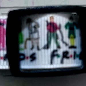Puede incluir: Cinta blanca con una hebilla negra y una impresión colorida de los seis personajes del programa de televisión "Friends". La cinta tiene el texto "Friends" impreso en ella.