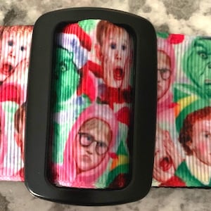 Puede incluir: Una correa de guitarra negra con fondo blanco que presenta un patrón repetido de varios personajes de la película 'Elf'.
