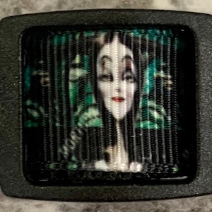 May include: Black and green dog collar with a repeating pattern of a woman with long black hair and a green dress. The collar has a black buckle with a square image of the woman in the center.