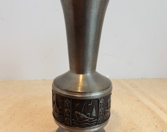May include: A silver-colored metal vase with a flared top and a round base. The vase features a detailed band of embossed sailboats around the middle. The vase is likely made of pewter or a similar metal.