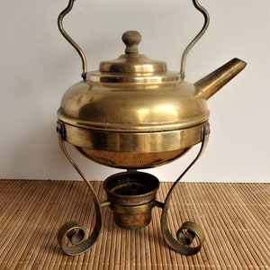May include: A vintage brass tea kettle with a black handle and wooden knob. The kettle sits on a matching brass stand with a burner. The stand has decorative scrollwork. The kettle is designed to keep tea warm.