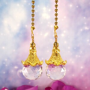 May include: Two gold-colored ceiling fan pull chains with clear crystal ball pendants. The pendants have a gold-colored textured cap. The background is a soft blend of blue, purple, and pink with a sparkling effect.