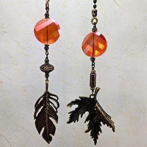May include: Two decorative ceiling fan pulls. Each pull features a faceted orange bead, a decorative metal piece, and a bronze-colored leaf or feather charm. The pulls hang from a dark chain.