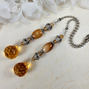 May include: Two decorative ceiling fan pull chains. Each chain features a faceted amber-colored crystal teardrop, brown oval beads, and silver-toned metal accents. A silver ball chain connects the two pulls.