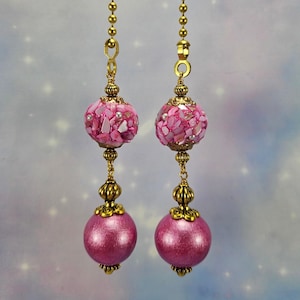 May include: Two decorative ceiling fan pulls. The top orbs are covered in pink shell chips and small pearls. The bottom orbs are a solid, shiny pink. The pulls are connected by gold-colored chains and decorative filigree.